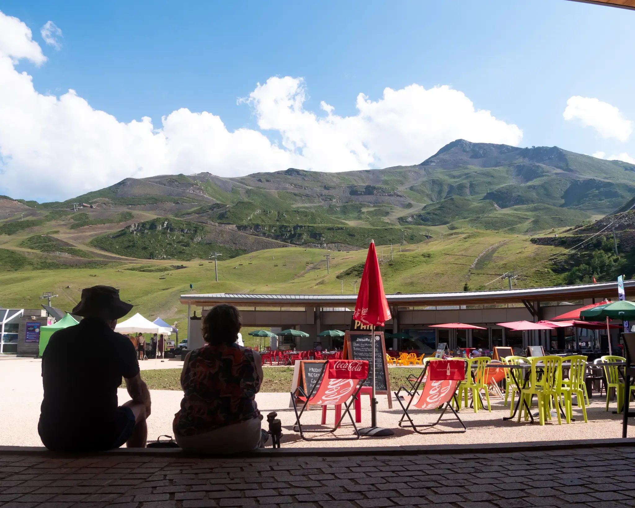 Deux personnes de dos observent la terrasse d'une station de montagne face à de hauts sommets verdoyants sous un ciel bleu.